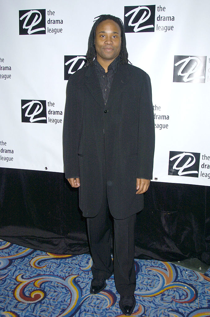 Billy in a black suit stands in front of a Drama League backdrop