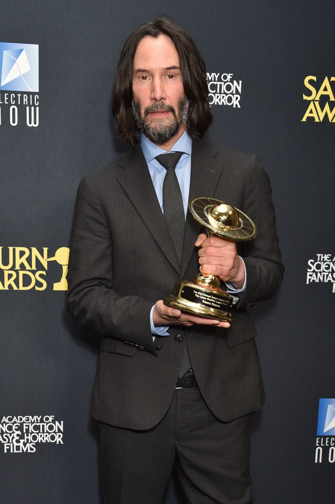 Keanu Reeves with facial hair and longish hair in a classic suit and tie holding a Saturn Award trophy