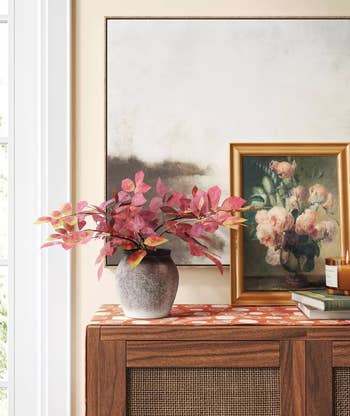 Elegant home decor setting with a vase of pink leaves on a wooden cabinet beside a framed painting and candles