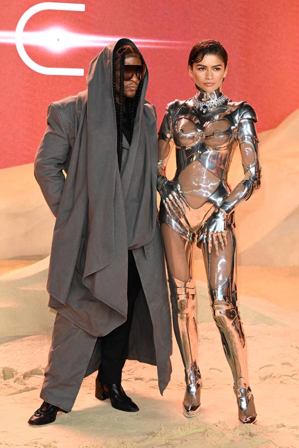 Law Roach and Zendaya on red carpet, one in oversized gray suit and hood, the other in metallic bodysuit with structured shoulders
