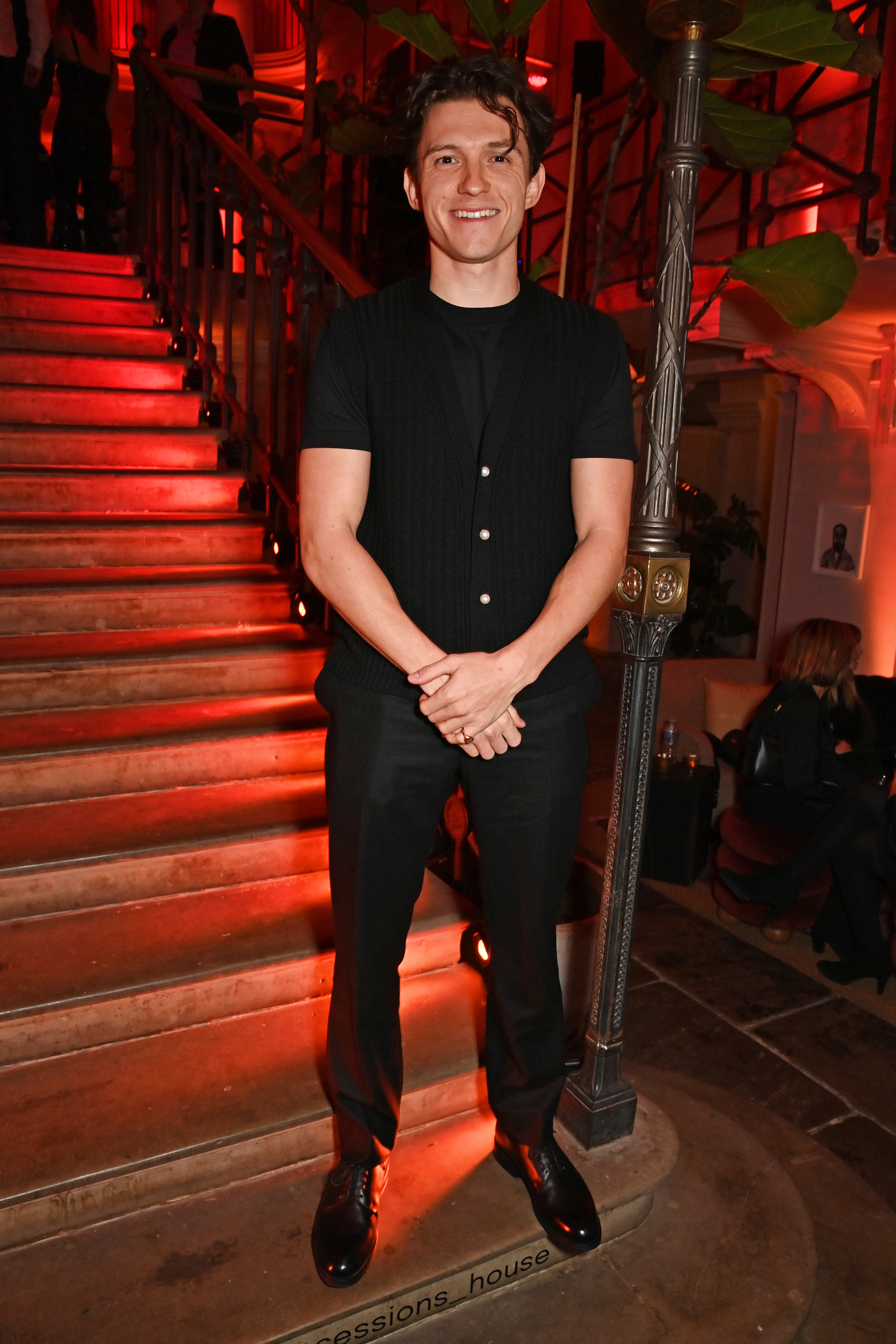 Tom Holland standing on stairs at an event, wearing a short-sleeve black shirt and trousers with shiny black shoes