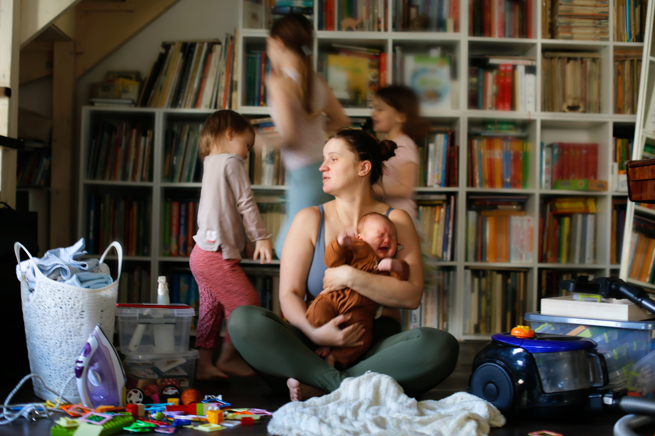 A mother holds a baby and sits among toys as another child and adult move in the background, depicting a lively family scene