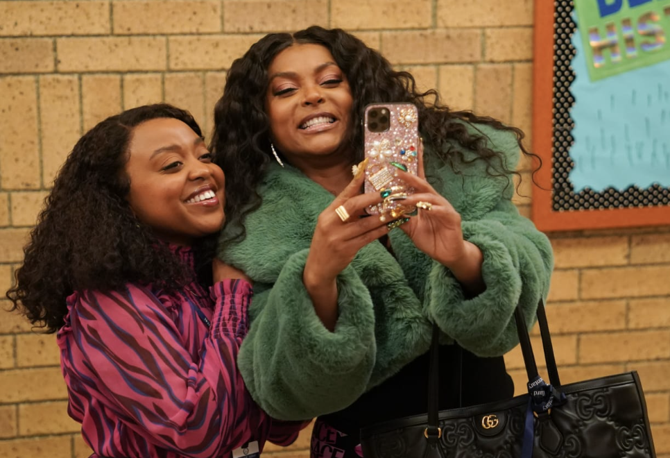 Two women smiling, taking a selfie with a bedazzled phone case. They are indoors, standing close, showing a friendly pose
