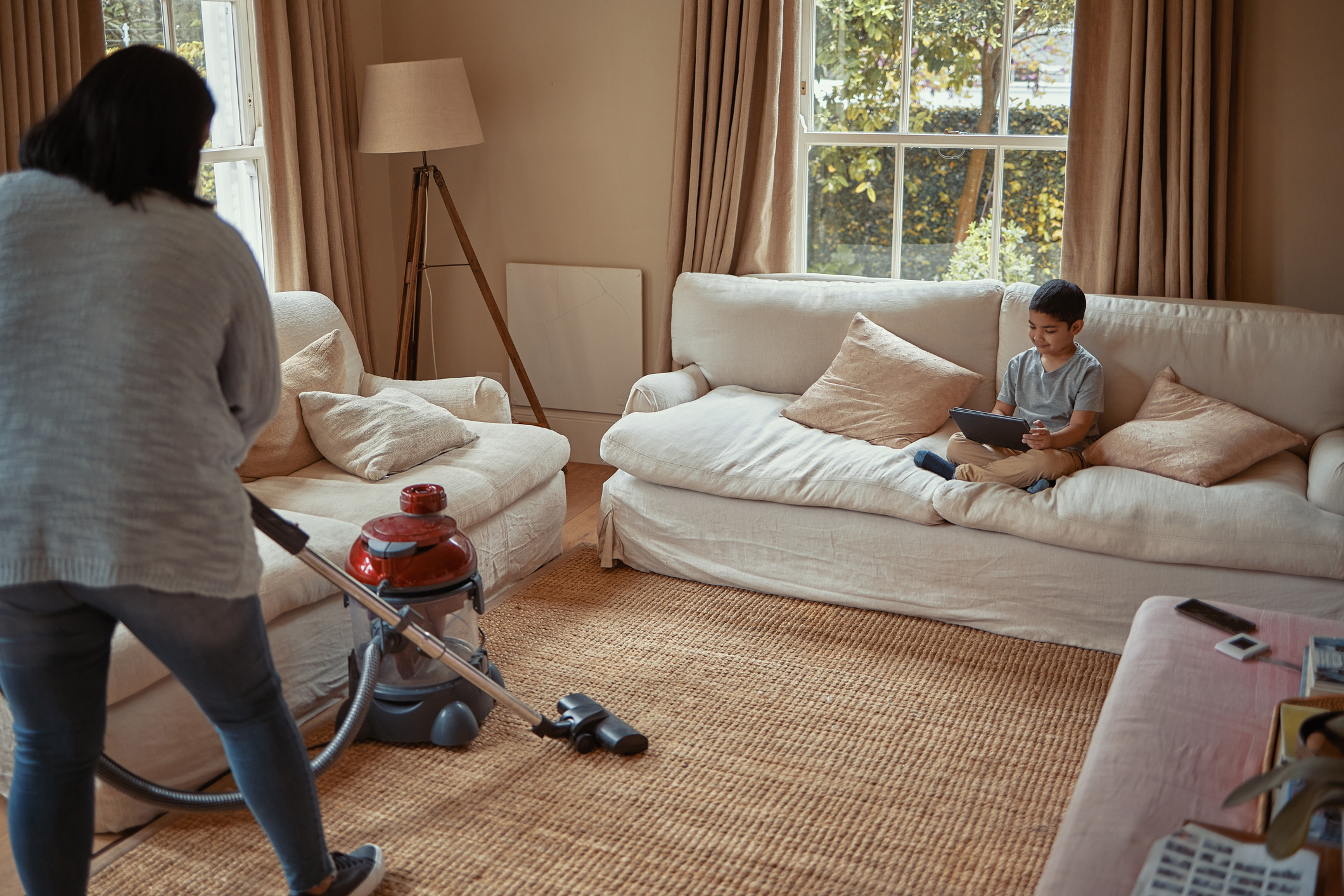 Mom vacuums while child sits on a sofa with a tablet in a living room setting