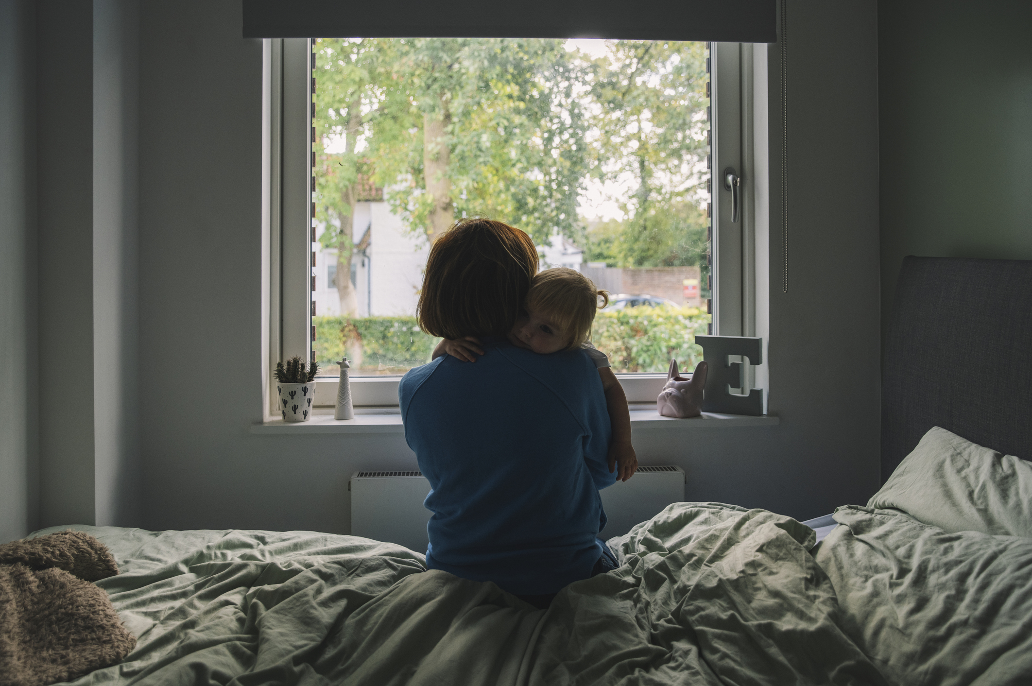 Mom and child embracing, looking out a window from a bed, in a dim bedroom