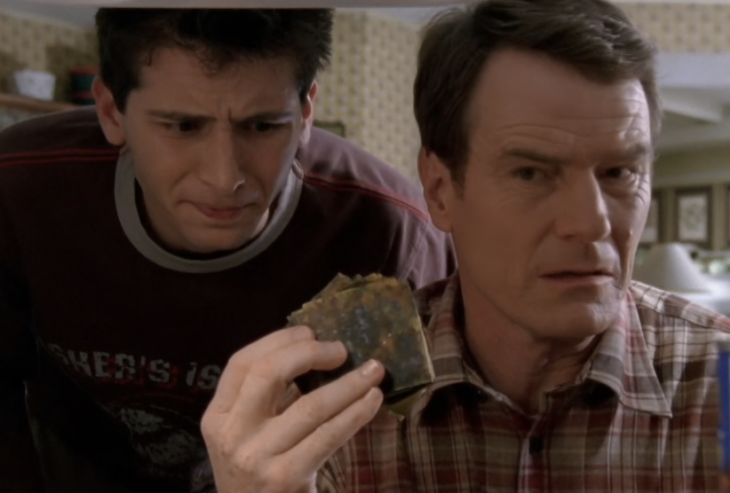 Two TV show characters, a young man and an older man, look concerned at a burned item the older man holds