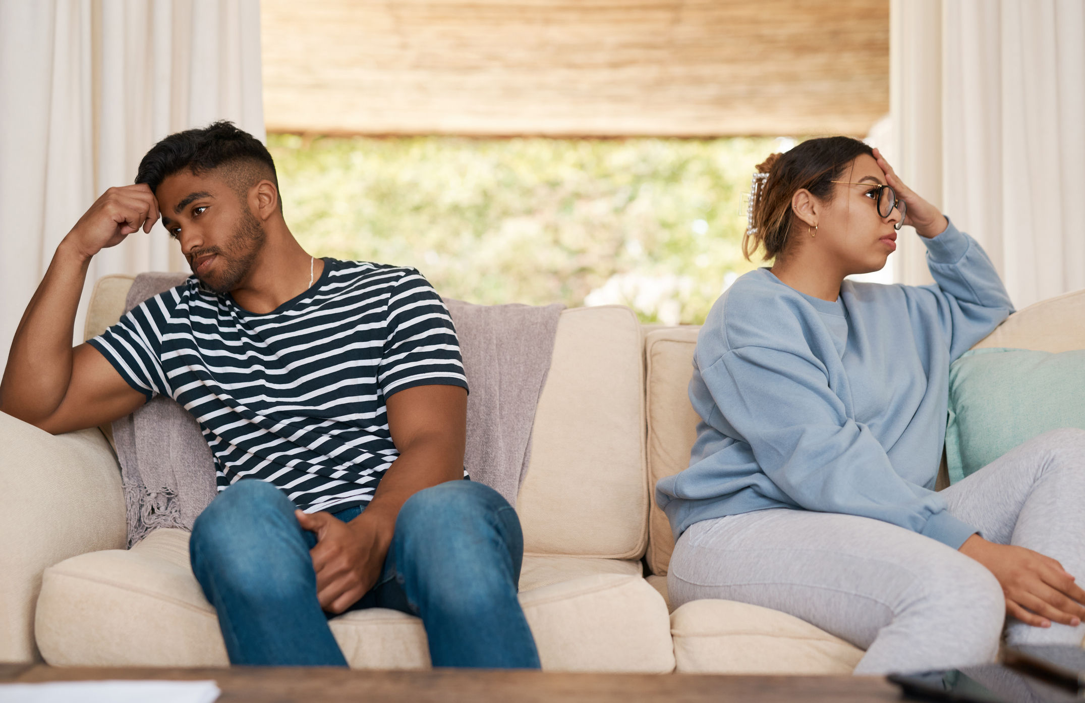 An upset couple sit on a couch not facing each other, both appearing thoughtful and distant