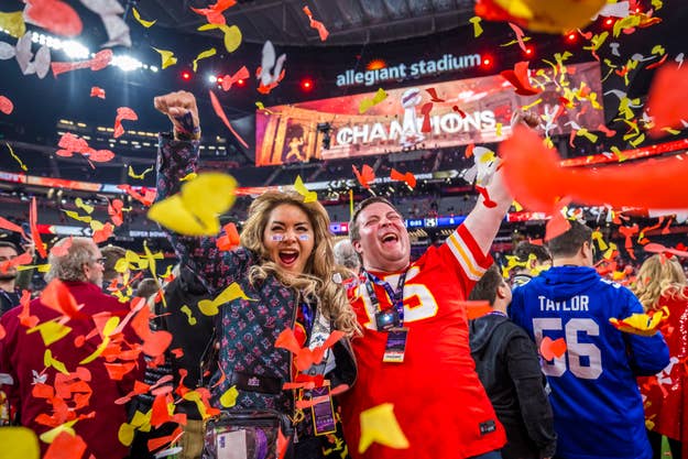 Two excited fans celebrate amidst falling confetti at a stadium with a "CHAMPIONS" sign in the background