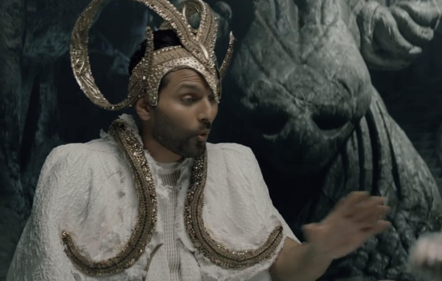 Jay in a detailed costume with headdress sits beside a CGI creature, expressing surprise or conversation