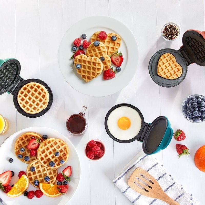 mini waffle makers on a table with plates of waffles, fruit, and a fried egg, suggesting a variety of breakfast options
