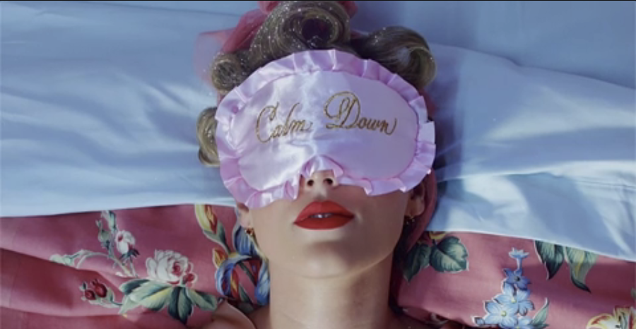 Person lying down with an eye mask that reads &quot;Calm Down&quot;, red lips, on a bed