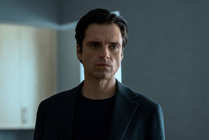 Man in a dark jacket stands indoors with a serious expression