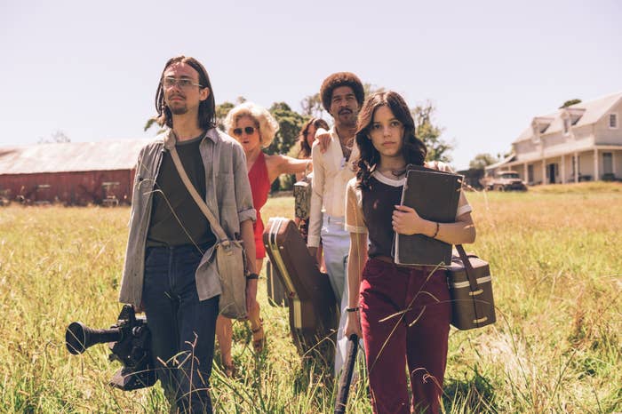 Four actors pose dramatically in an open field, dressed in 70s-inspired costumes, with film equipment