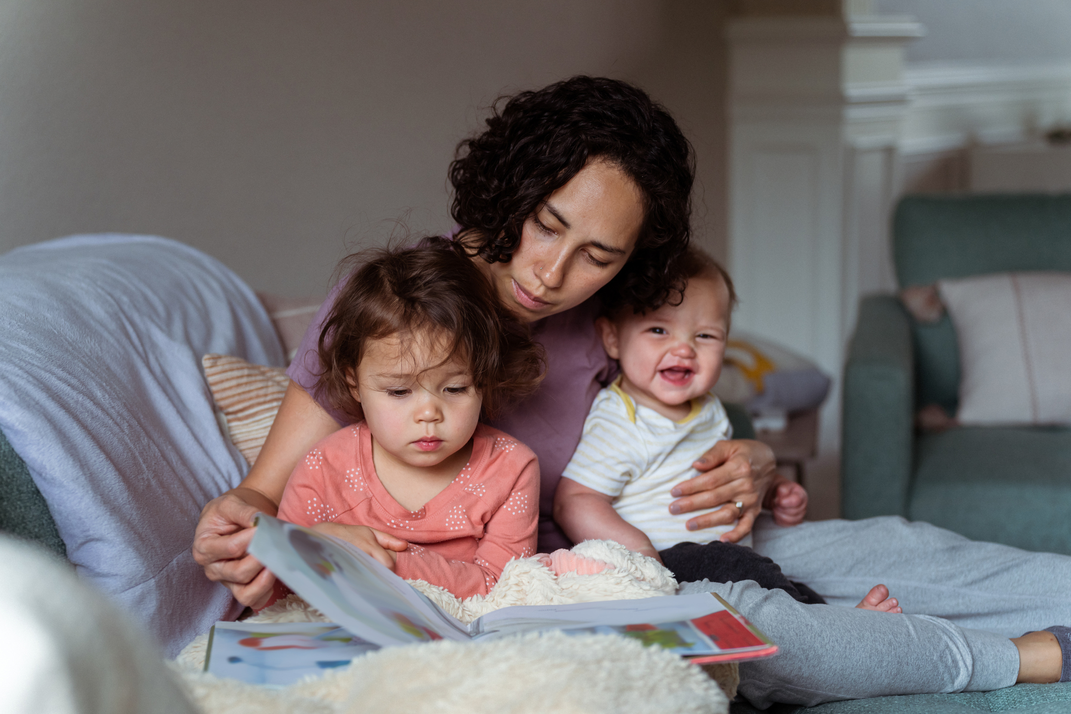 Woman reads book with toddler and baby smiling on bed