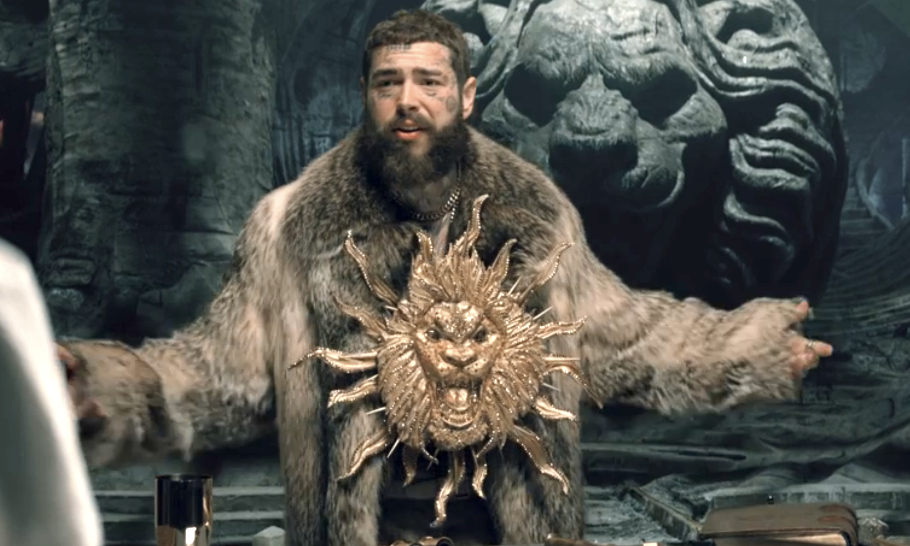 Post Malone in elaborate costume with a lion-faced chest piece, thick beard, and fur cloak, gesturing widely in a stone room