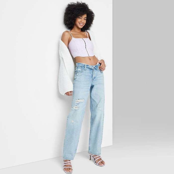 Woman in a stylish crop top, torn jeans, and white cardigan posing with a smile
