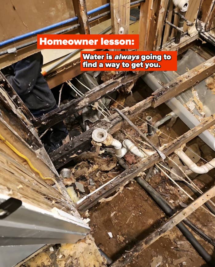 A damaged bathroom floor with exposed pipes and joists, and a person's legs visible