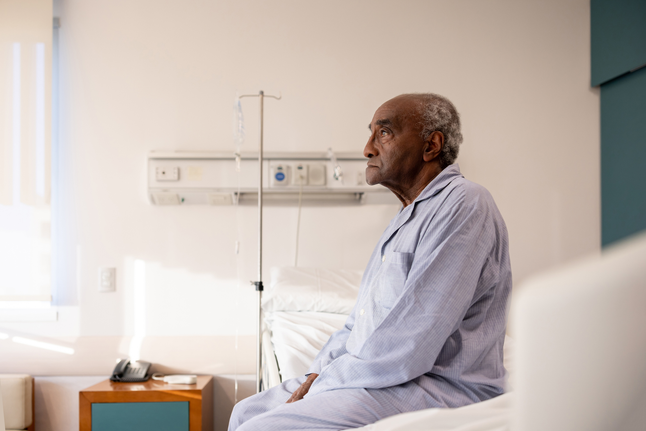 Older man in a hospital gown seated on a bed, looking thoughtful, in a hospital room