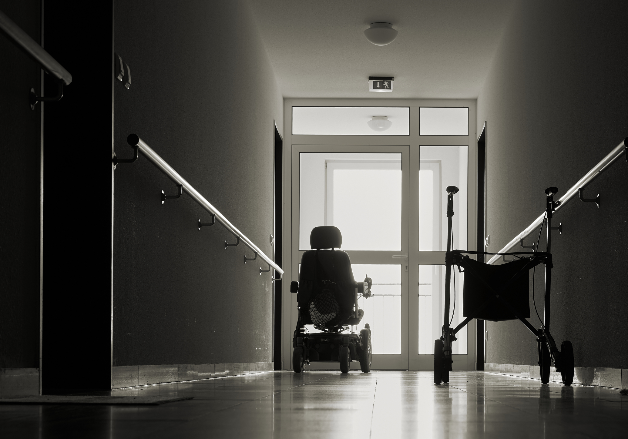 Person in a wheelchair at the end of a dimly lit hallway, facing a bright doorway, with a walker on the right side