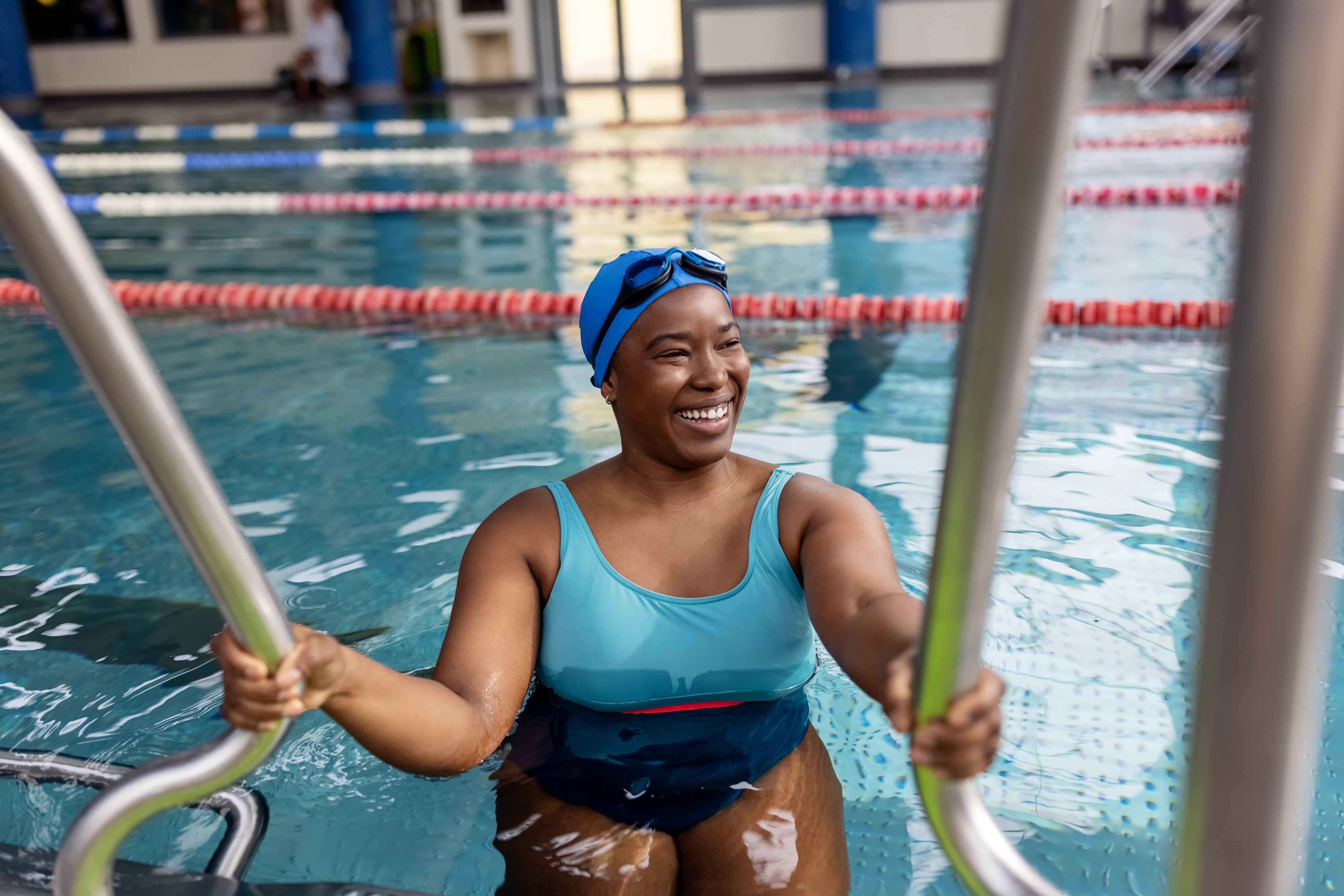 Woman in a swimsuit with a swim cap and goggles, smiling, holding onto a pool ladder