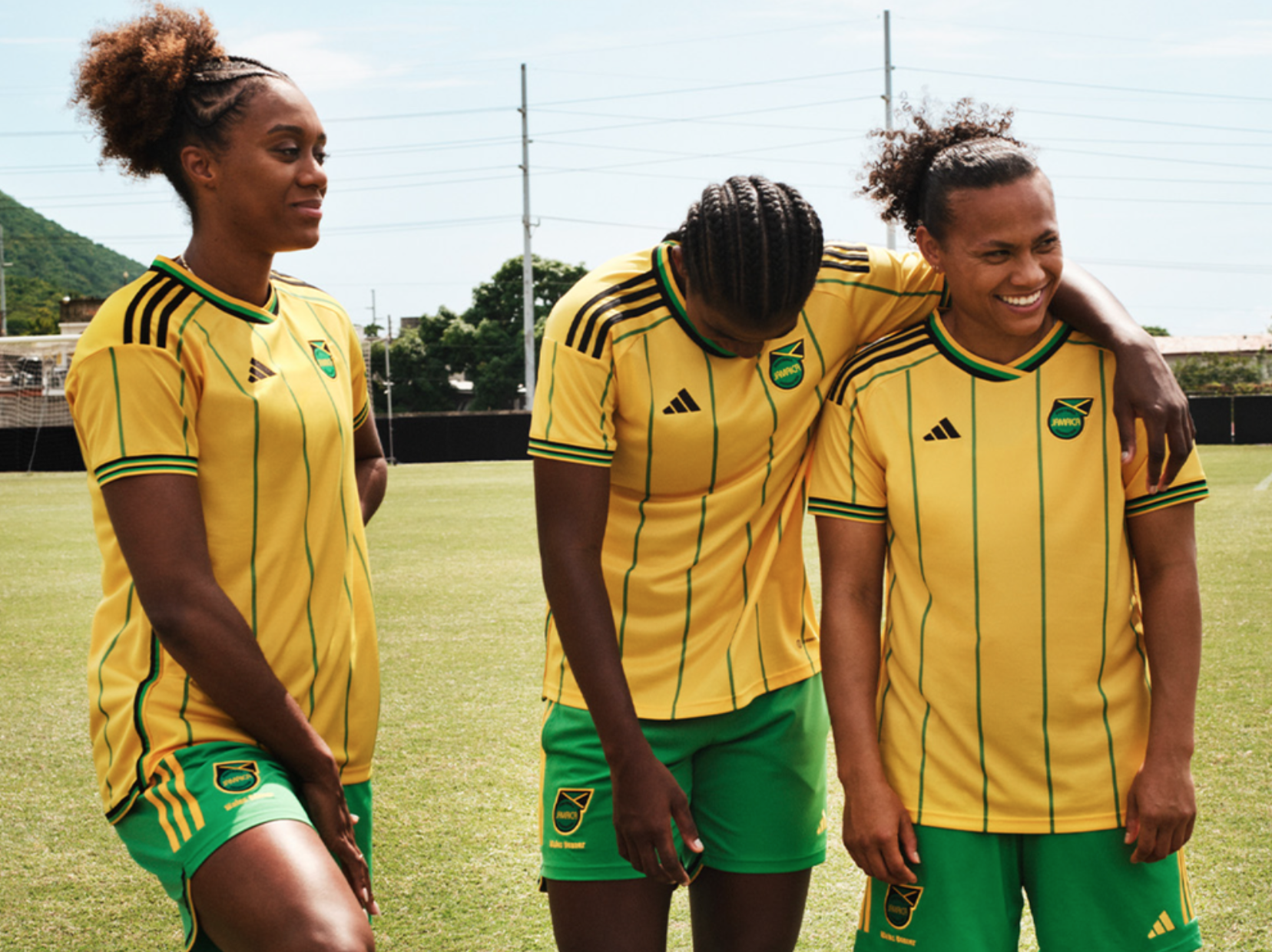 Three soccer players in yellow and green jerseys, one looking down at another&#x27;s sneakers, on a field