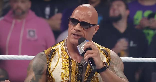 Dwayne Johnson in a wrestling ring speaking into a microphone while wearing a black and gold outfit