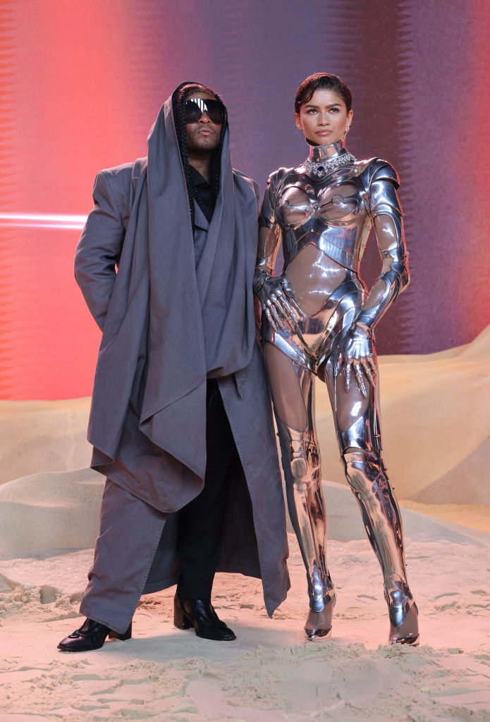 Kanye West in a layered gray outfit stands next to Kim Kardashian in a metallic bodysuit at an event