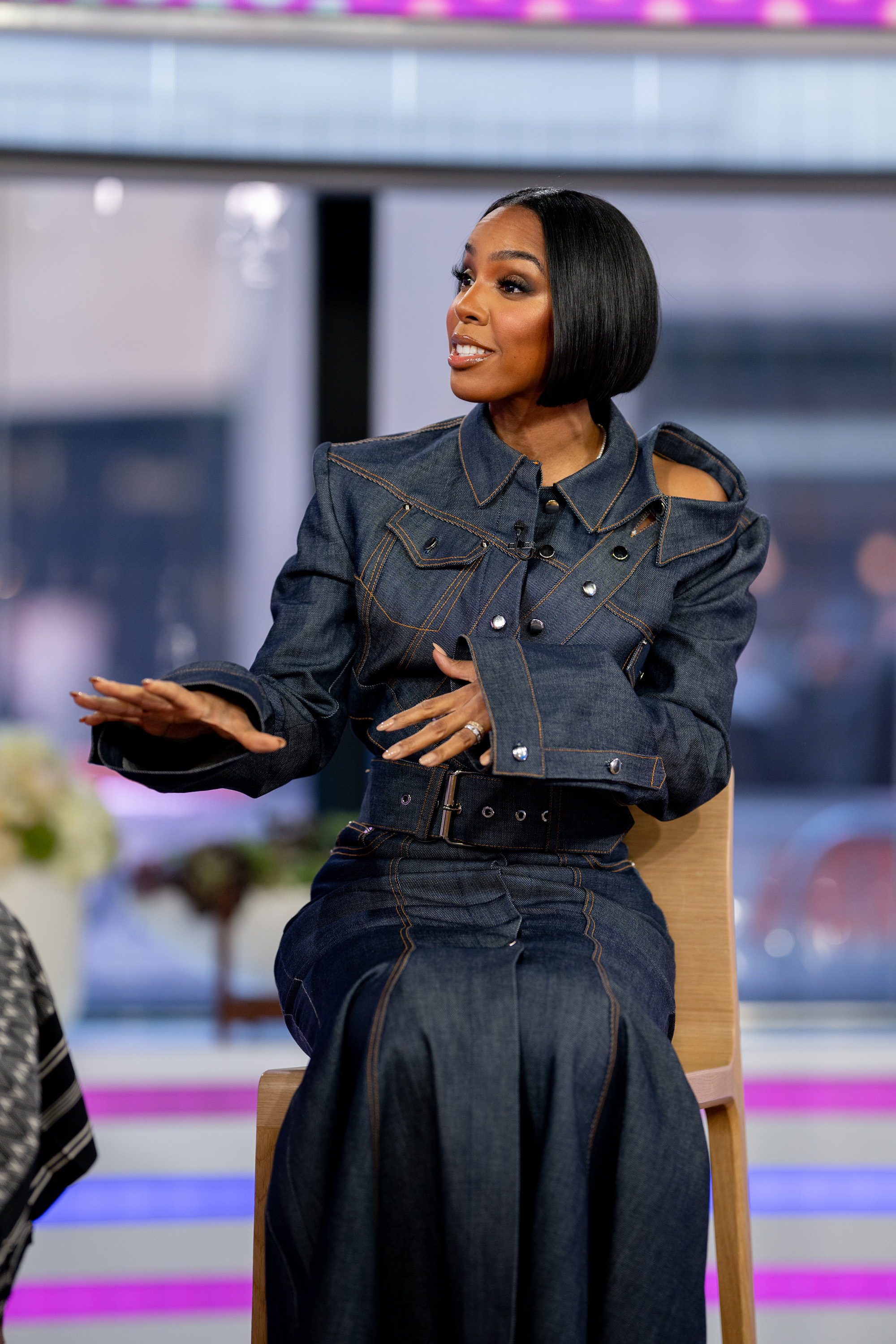 kelly in a stylized denim jacket and skirt speaks while seated, gesturing with her hand