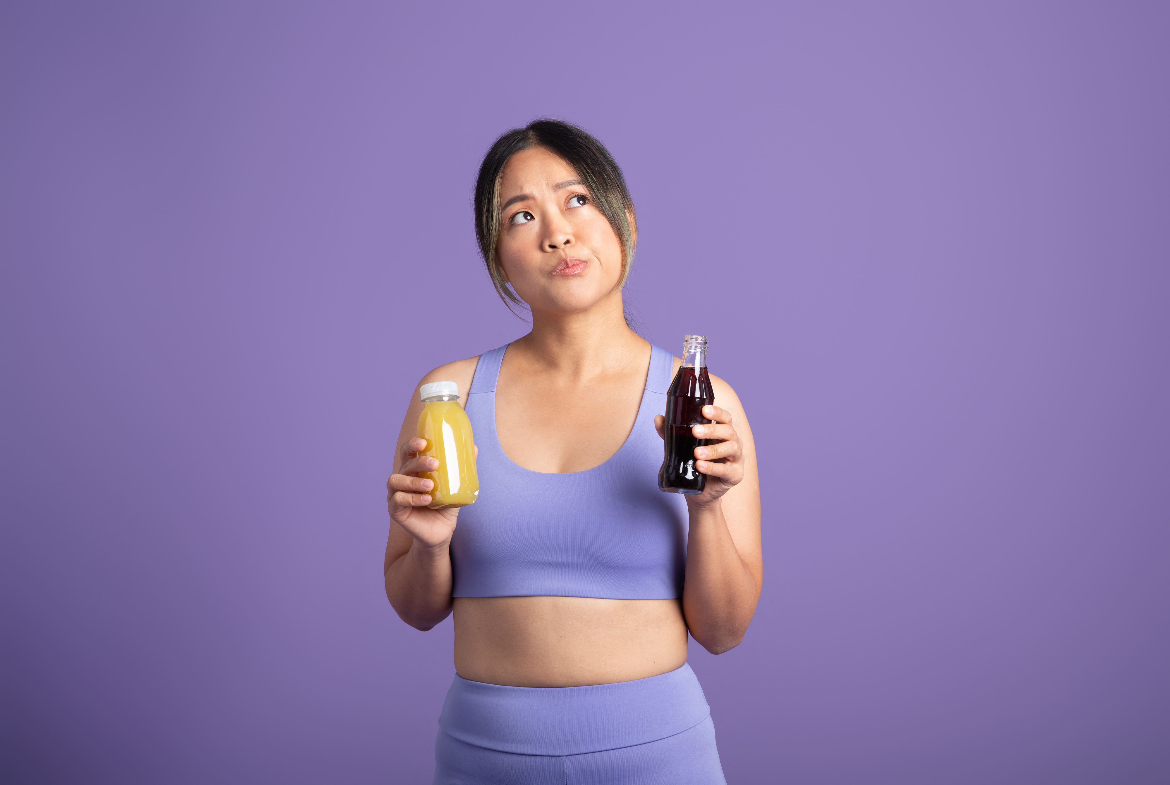 Woman in sportswear holding a juice bottle and a soda, looking contemplative