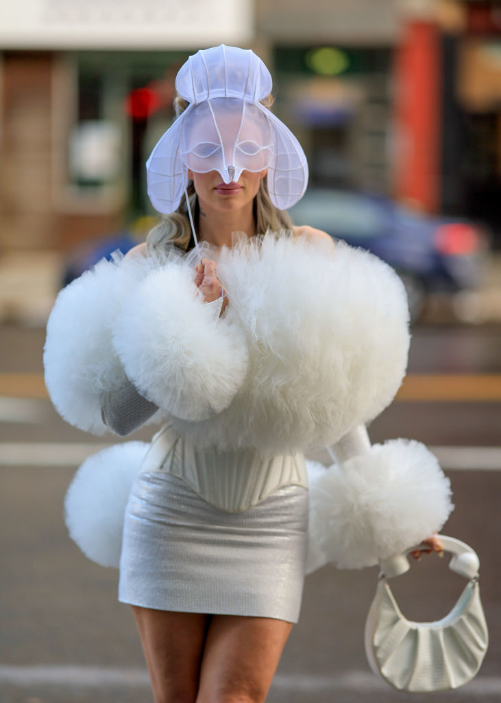 Julia in a white fluffy dress with matching hat, mask, and handbag on a city street