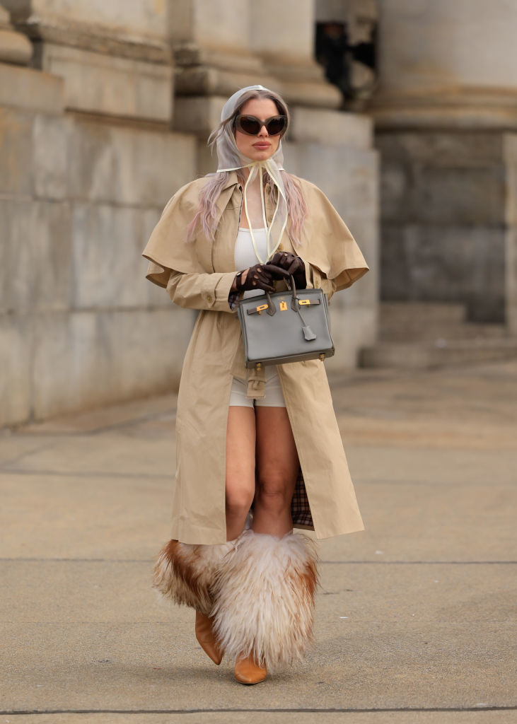 Julia in trench coat, fur boots, holding bag, sunglasses, and headscarf