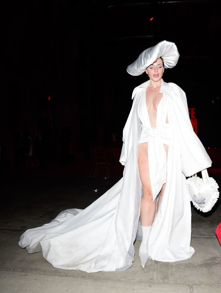 Julia in an extravagant white outfit with a large hat, cape, and high slit, at an event