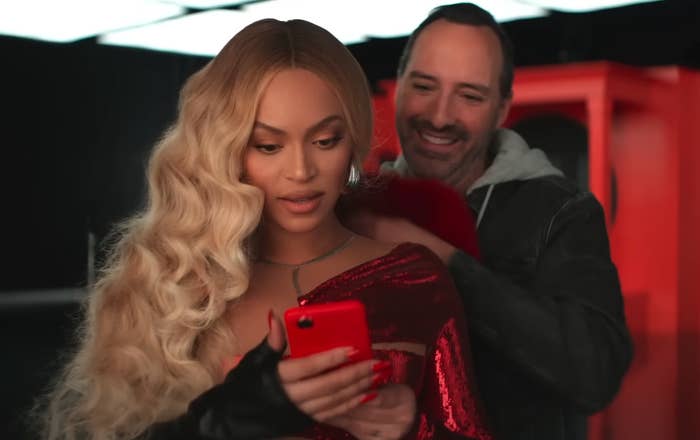 Beyoncé in a glittery outfit, looking at a phone, with a smiling man in a hoodie behind her