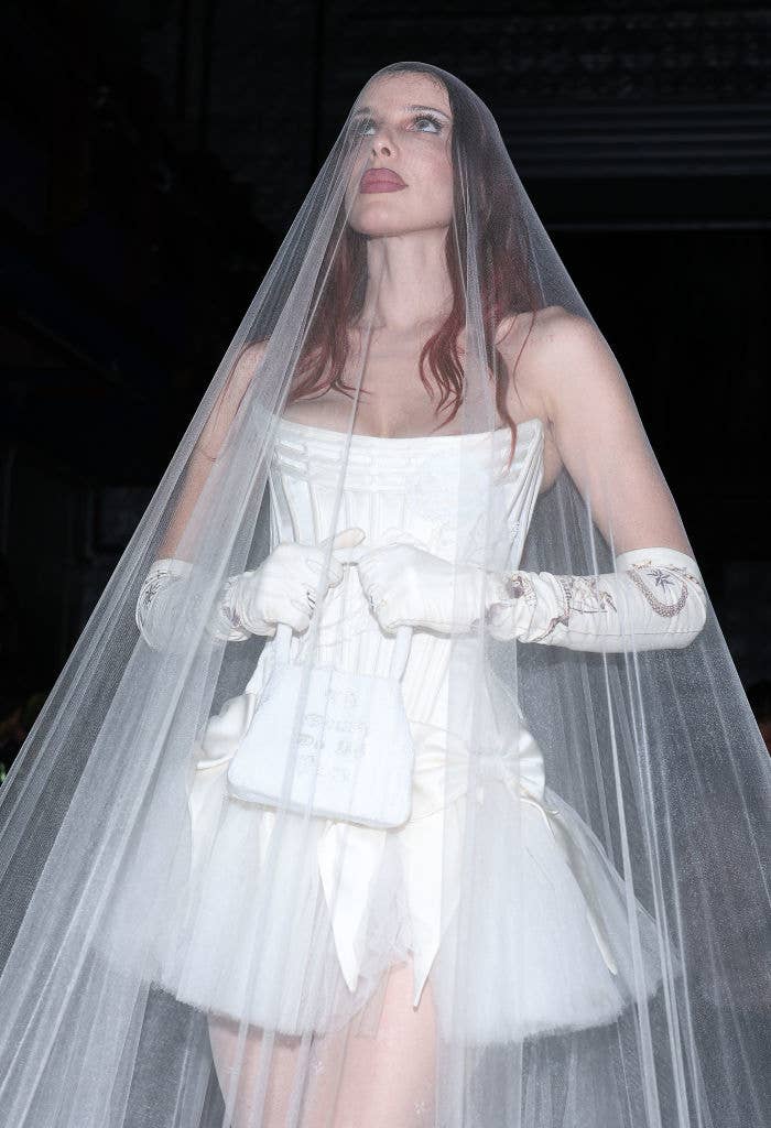 Julia in a wedding dress and veil holding a white purse, gloves on hands, looking upwards