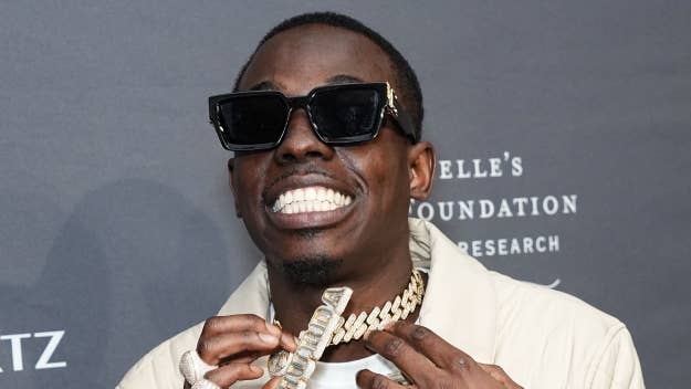 Bobby Shmurda in sunglasses and cream blazer grinning, holding chain necklace, at music event