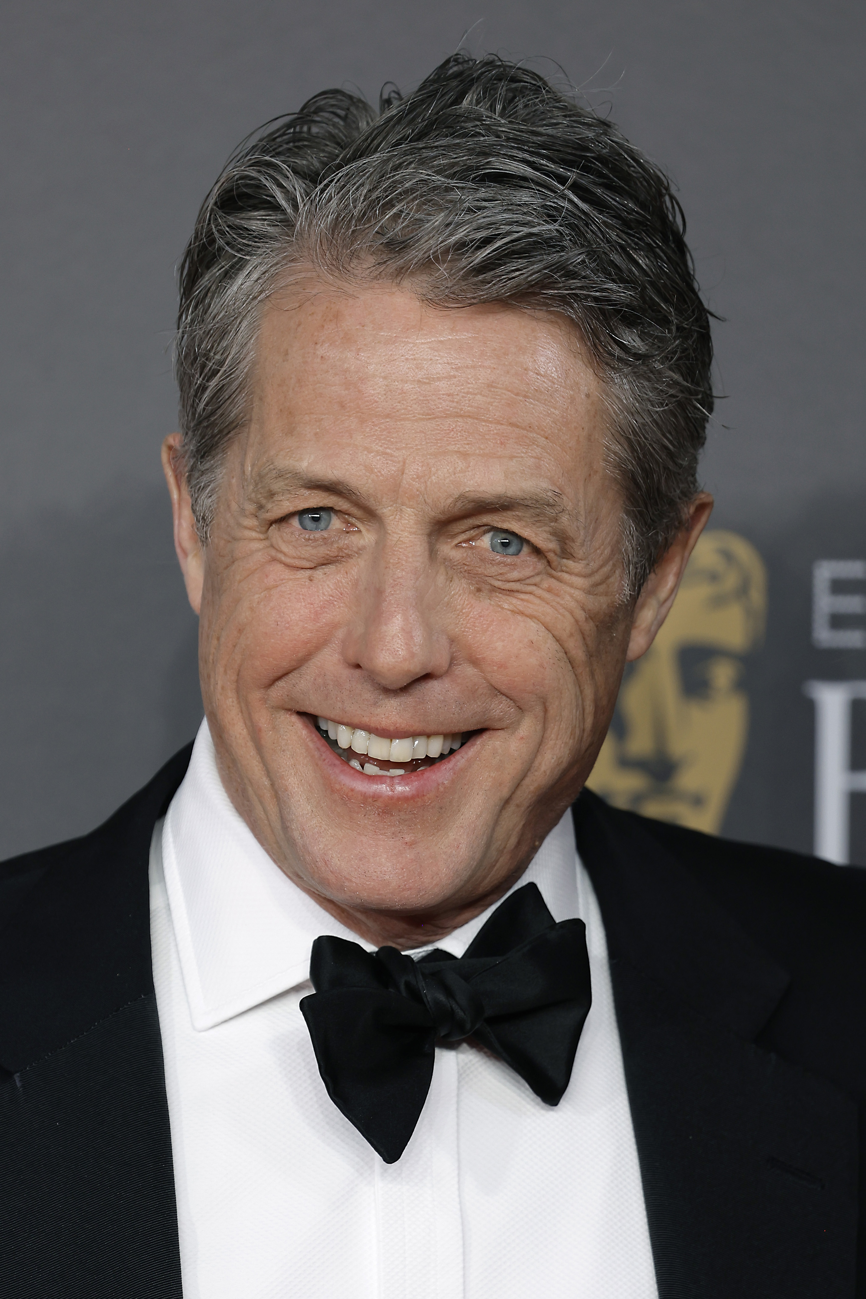 Hugh Grant smiles in a black tuxedo with a bow tie at an event