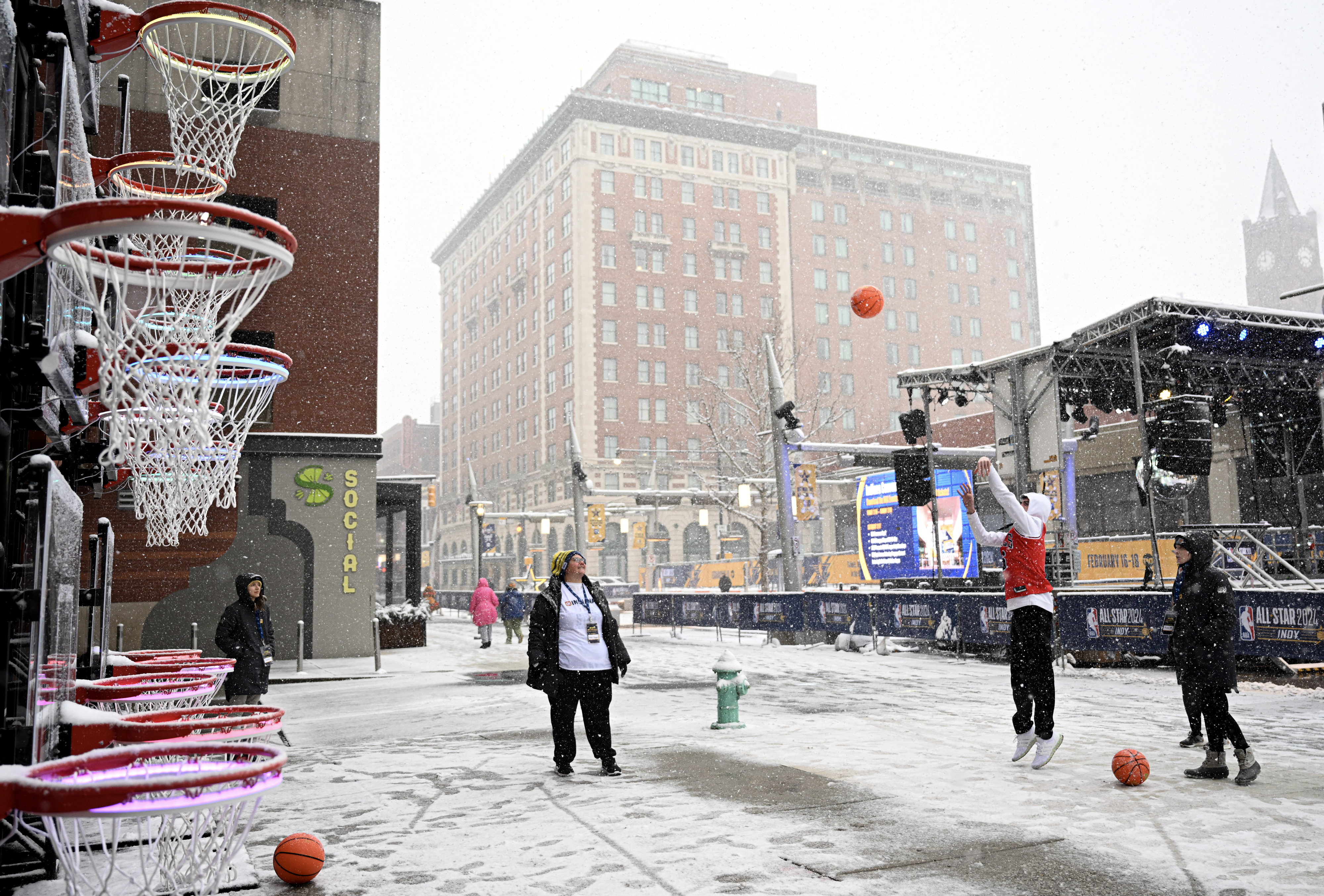 Basketball hoops line a snowy street where people are playing, with buildings and a large digital display in the background