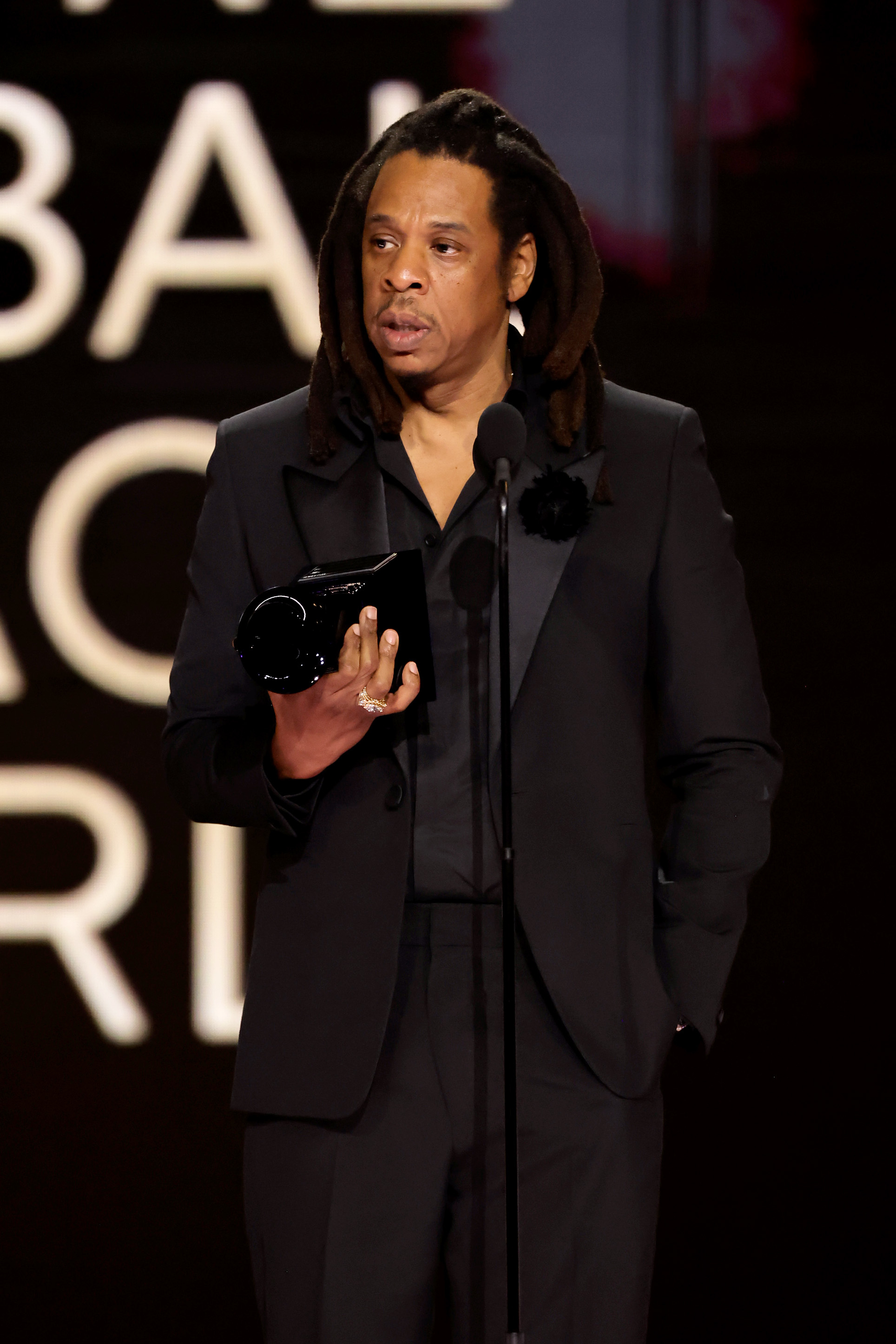 Jay-Z speaks at a podium during an awards ceremony, wearing a formal black suit and holding a trophy