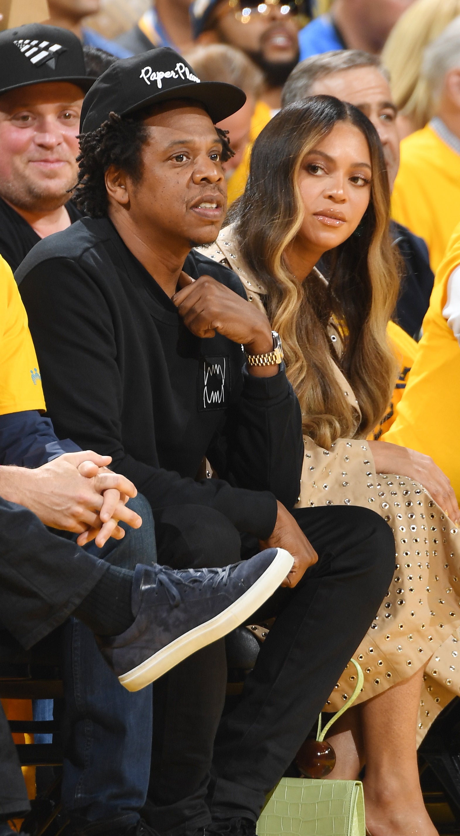 Jay-Z wearing a black sweatshirt and cap seated courtside with Beyoncé in a dotted dress and heels