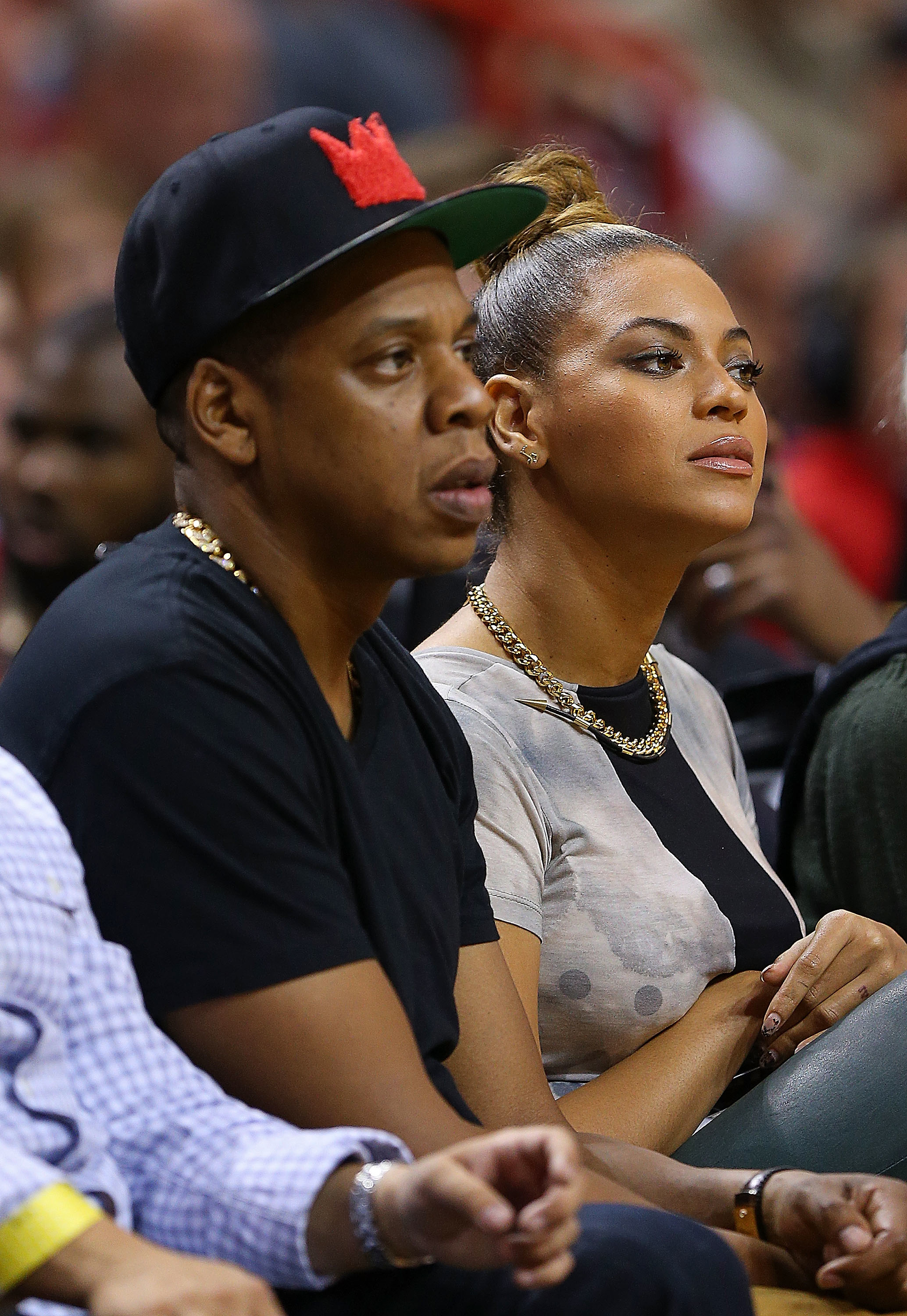 Jay-Z and Beyoncé sitting courtside at a basketball game. Jay-Z wears a black cap and Beyoncé sports a chain necklace