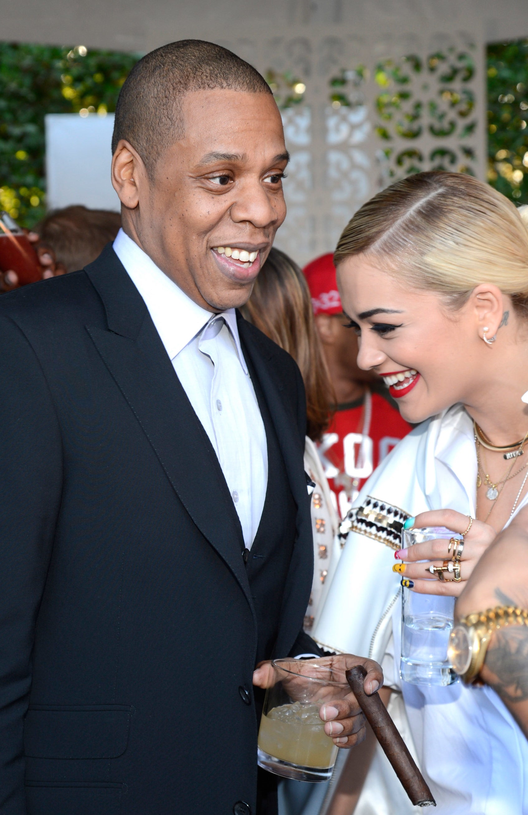 Jay-Z in a black suit seen conversing with Rita Ora in a patterned outfit, both smiling