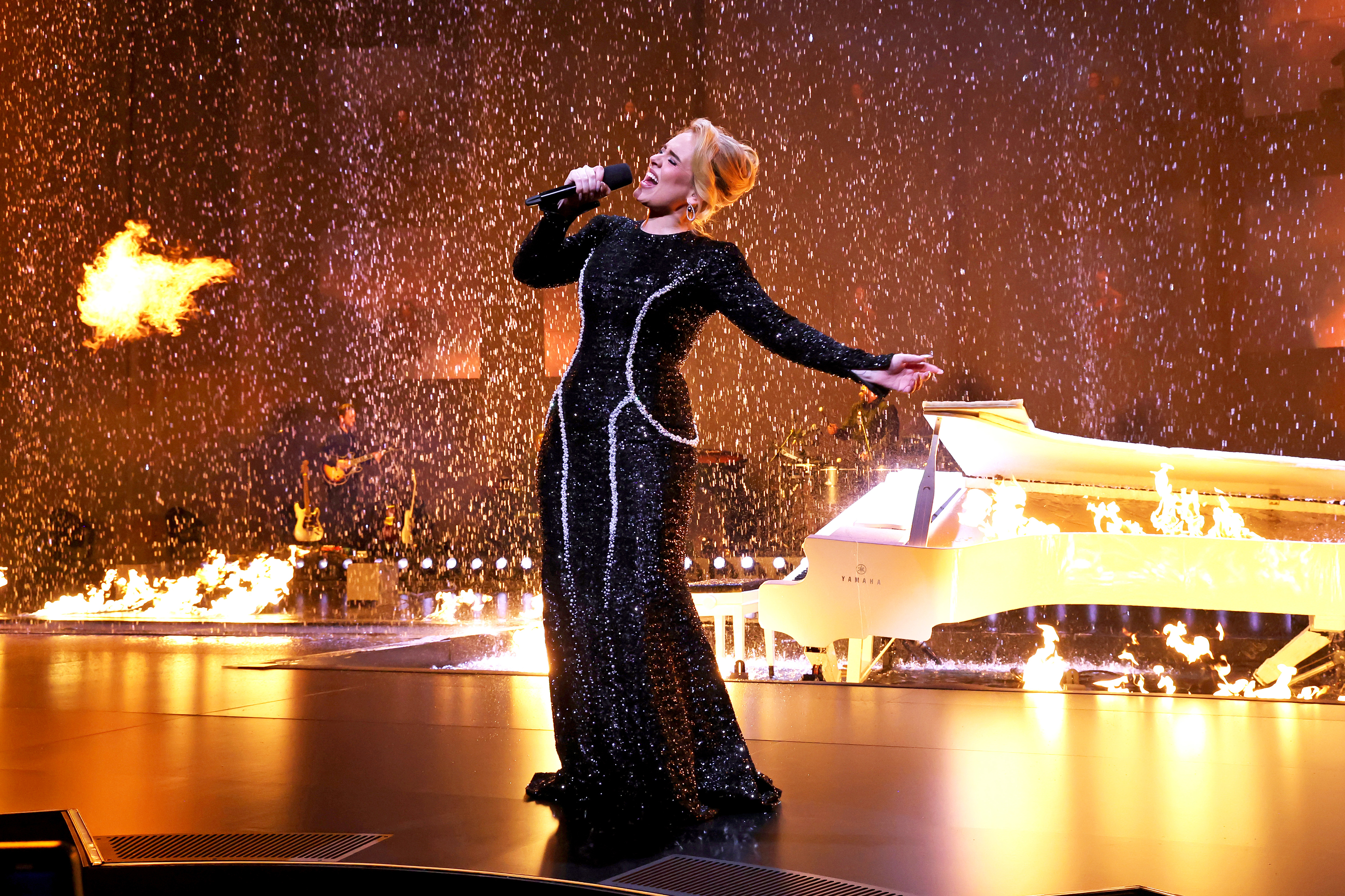 Adele performing on stage in a black long-sleeve gown with a sparkling neckline, near a white grand piano and pyrotechnics