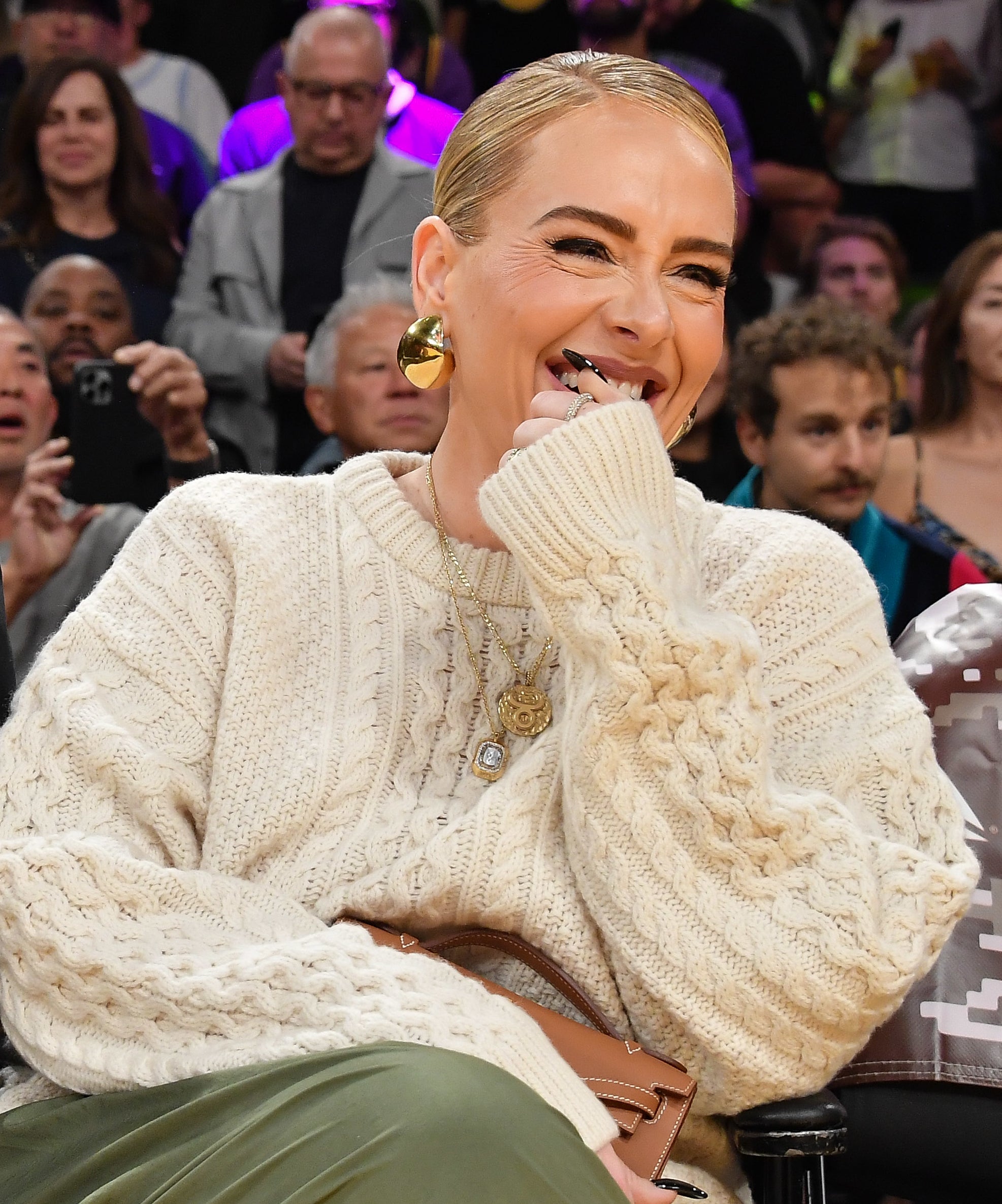 Adele in a knitted sweater and trousers, laughing with hand near mouth, sitting courtside