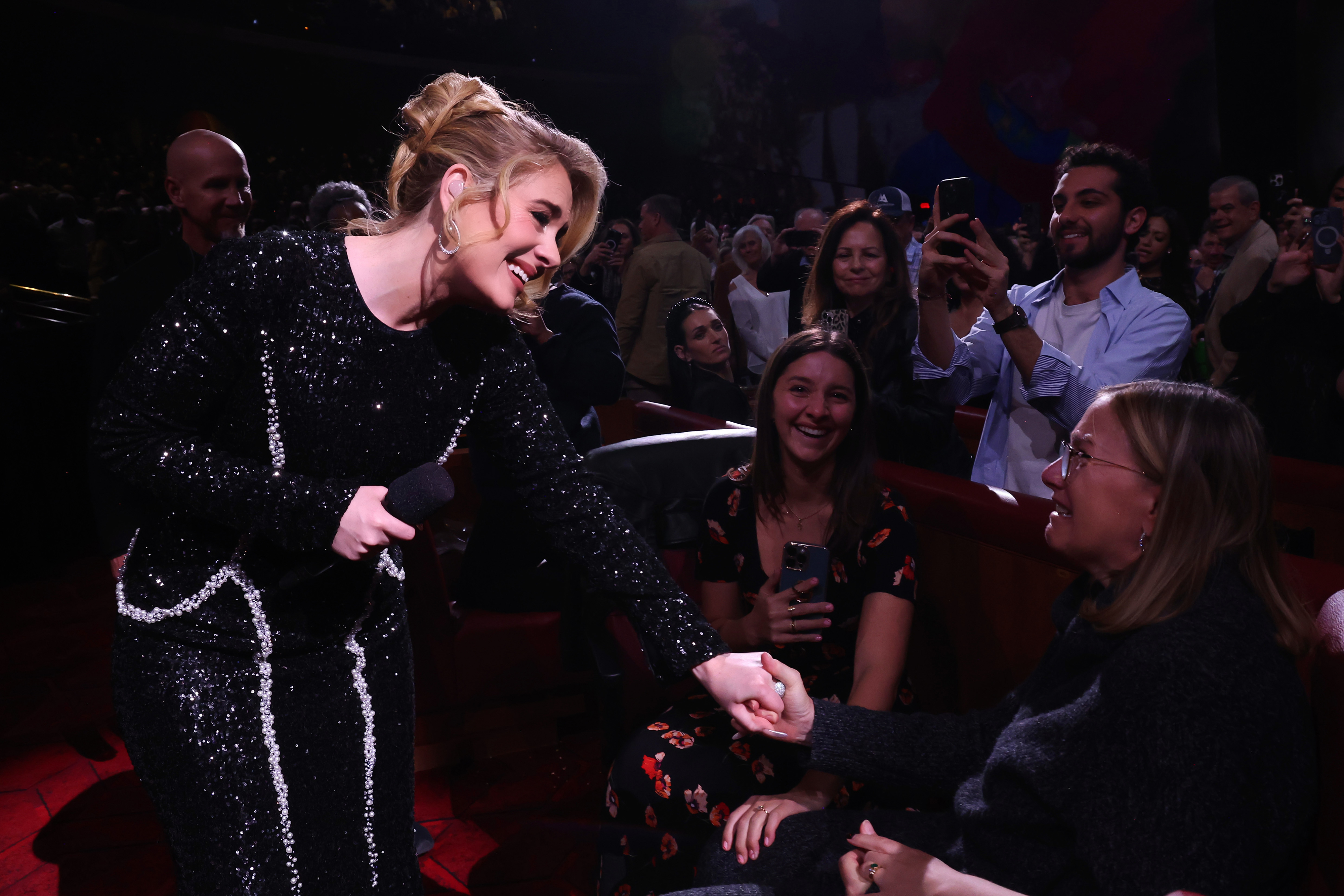 Adele shaking hands with a fan, wearing a sparkling outfit, at an indoor event surrounded by an audience