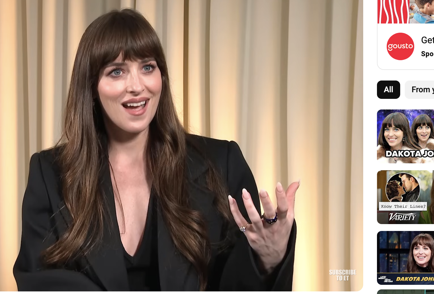 Dakota Johnson in an interview, gesturing with her hands, seated indoors with a plant and TV screen in the background
