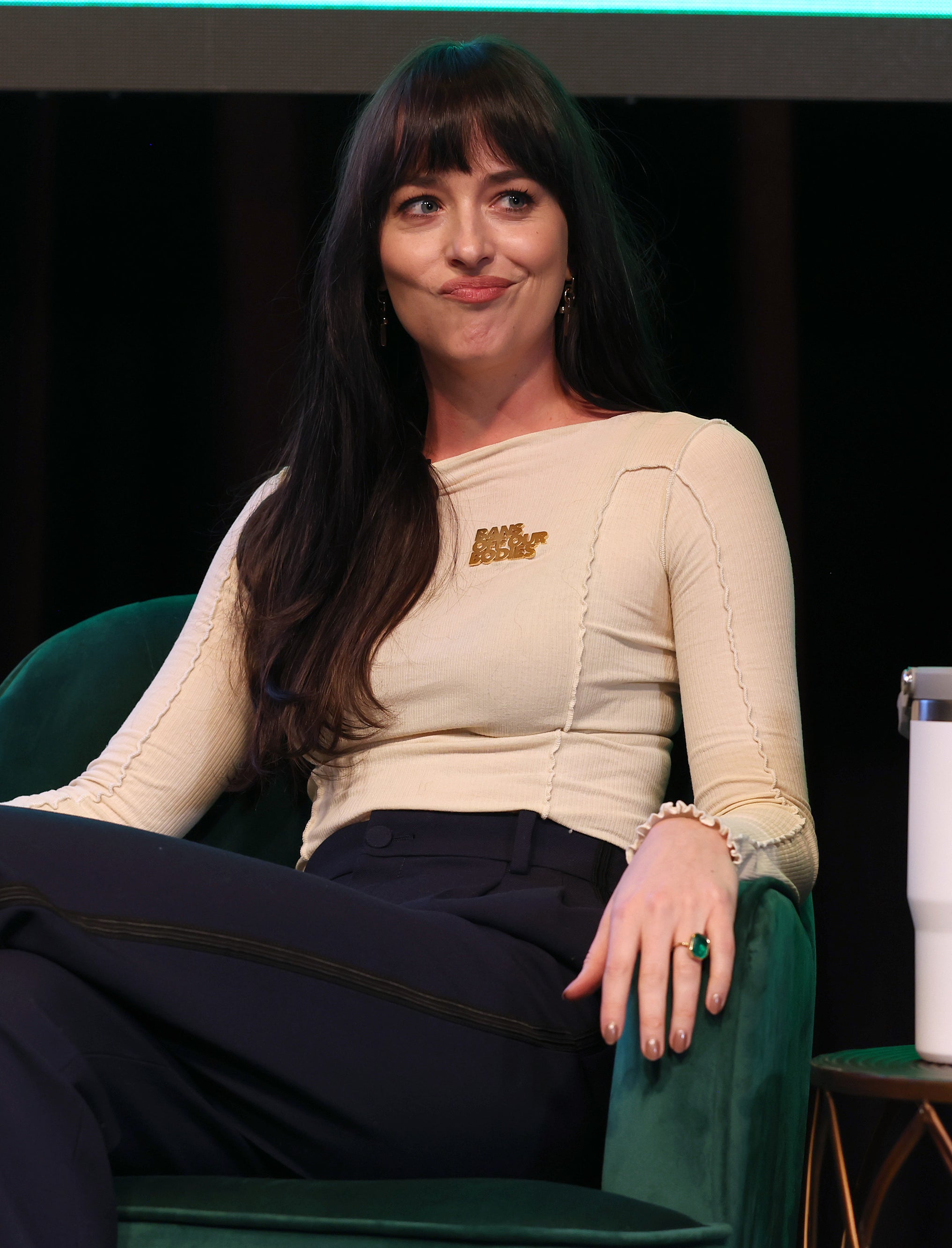 Woman seated on stage with a tumbler beside her, wearing a casual top and trousers