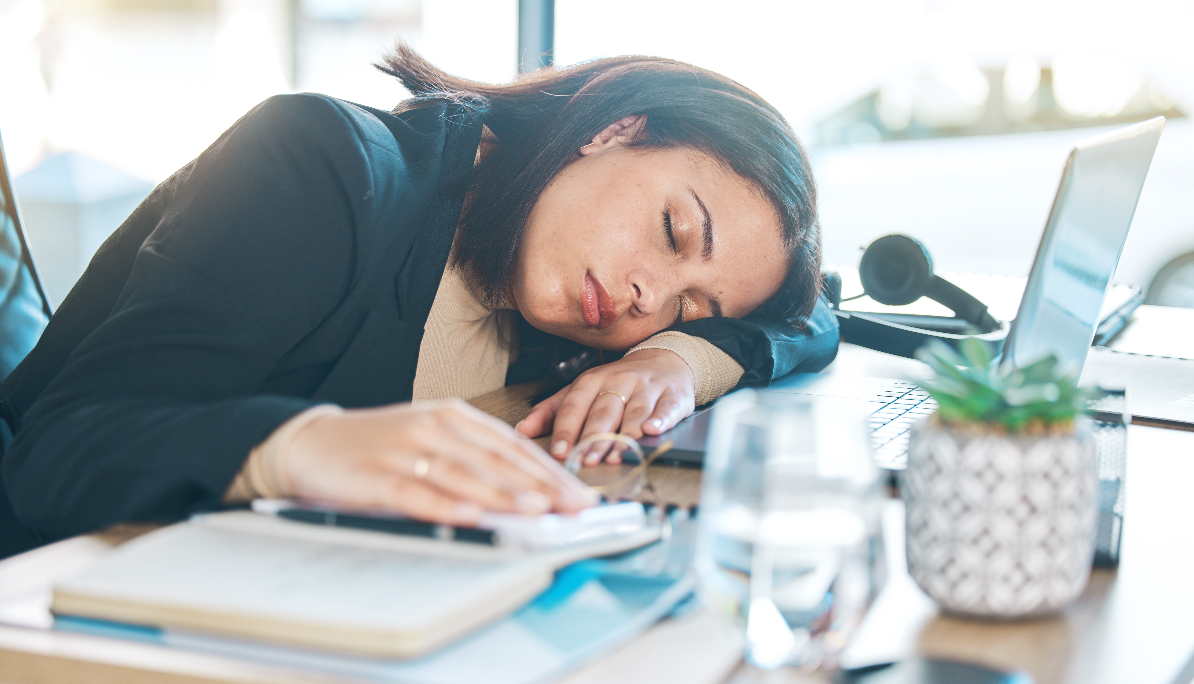 Woman asleep at desk with laptop, paperwork, and a plant, suggesting work fatigue or burnout