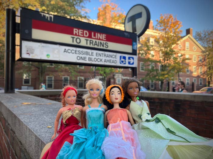 Four Barbie dolls wearing red, aqua, orange, and green dresses, sitting near the Harvard MBTA station in Cambridge, Massachusetts.