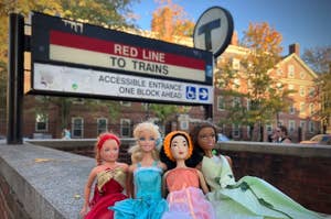 Four Barbie dolls wearing red, aqua, orange, and green dresses, sitting near the Harvard MBTA station in Cambridge, Massachusetts.