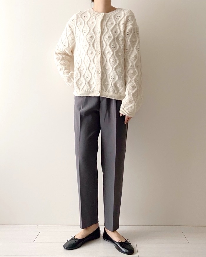 Person wearing a cable knit sweater and gray trousers with black flats, standing against a neutral background